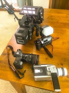A variety of cameras are placed on a wooden table, including a large video camera with a monitor, a DSLR with a zoom lens attached, an older film camera, and a vintage movie camera. The cameras are positioned together, showcasing different types of photography and video equipment.