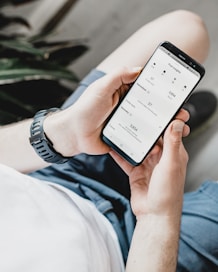 A person is holding a smartphone displaying an analytics or insights screen with various metrics and numbers. The individual is wearing a casual white shirt and a watch with a black band. The surrounding environment includes an indoor plant.