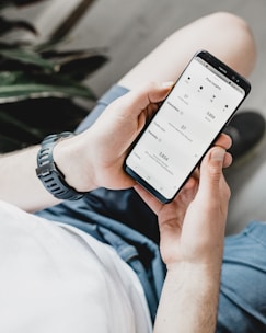 A person is holding a smartphone displaying an analytics or insights screen with various metrics and numbers. The individual is wearing a casual white shirt and a watch with a black band. The surrounding environment includes an indoor plant.