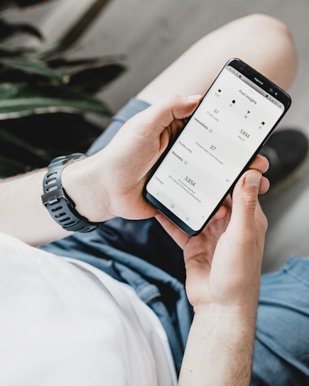 A person is holding a smartphone displaying an analytics or insights screen with various metrics and numbers. The individual is wearing a casual white shirt and a watch with a black band. The surrounding environment includes an indoor plant.