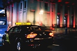 A taxi is driving down a city street at night, illuminated by streetlights and the brightly lit windows of a grand building in the background. The building is adorned with red lights, creating a striking visual effect. A blue bus is seen in the background.