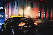 A taxi is driving down a city street at night, illuminated by streetlights and the brightly lit windows of a grand building in the background. The building is adorned with red lights, creating a striking visual effect. A blue bus is seen in the background.