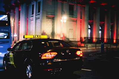 A taxi is driving down a city street at night, illuminated by streetlights and the brightly lit windows of a grand building in the background. The building is adorned with red lights, creating a striking visual effect. A blue bus is seen in the background.