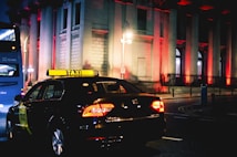 A taxi is driving down a city street at night, illuminated by streetlights and the brightly lit windows of a grand building in the background. The building is adorned with red lights, creating a striking visual effect. A blue bus is seen in the background.