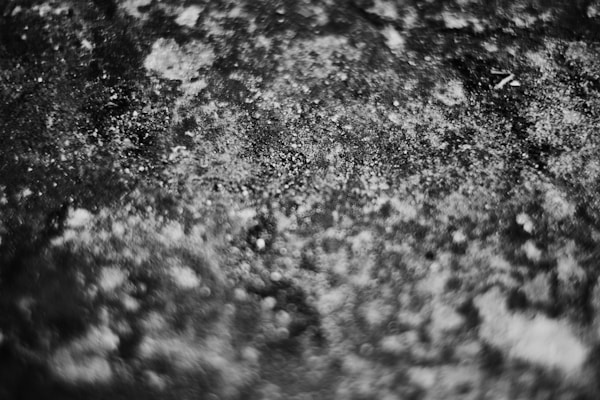 A black and white textured surface with various shades of gray. The texture appears rough and uneven, resembling stone or concrete with scattered white specks.