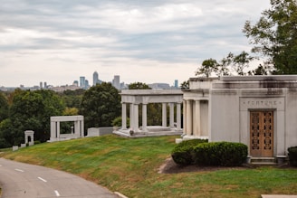 A tranquil cemetery setting with ornate mausoleums and gravestones surrounded by lush green grass and trees. A city skyline with tall buildings is visible in the distance under an overcast sky.