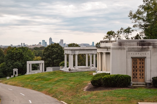 A tranquil cemetery setting with ornate mausoleums and gravestones surrounded by lush green grass and trees. A city skyline with tall buildings is visible in the distance under an overcast sky.