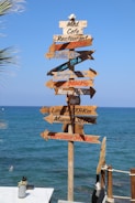 A wooden signpost with multiple directional arrows pointing to various cities and their distances is set against a backdrop of a clear blue sea. The names of cities like Rome, Beijing, Paris, Moscow, London, Miami, and Barcelona are inscribed on the arrows. A small plant and a rope fence are visible in the foreground, and the sky is cloudless.