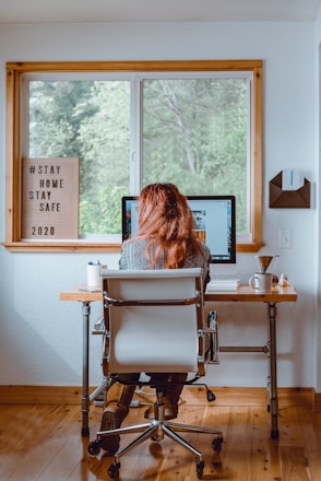 A person with long hair is sitting in an office chair at a wooden desk, working on a computer. The room has a large window overlooking green trees, and the walls are white. On the desk, there is a cup, a couple of pens in a holder, and some papers. A wooden letter board next to the window displays a message about staying home and safe. The environment appears homey and calm, conducive to focused work.
