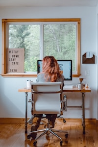 A person with long hair is sitting in an office chair at a wooden desk, working on a computer. The room has a large window overlooking green trees, and the walls are white. On the desk, there is a cup, a couple of pens in a holder, and some papers. A wooden letter board next to the window displays a message about staying home and safe. The environment appears homey and calm, conducive to focused work.