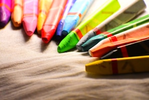 Multiple colorful wax crayons are lined up at the edge of a textured surface, displaying a variety of vibrant hues. The light highlights the texture and color details of the crayons, creating a vivid and artistic composition.