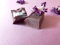 Two pieces of chocolate, one partially bitten, are garnished with dried purple flowers and placed on a pink textured surface. There are more dried flowers scattered in the background.