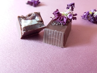 Two pieces of chocolate, one partially bitten, are garnished with dried purple flowers and placed on a pink textured surface. There are more dried flowers scattered in the background.
