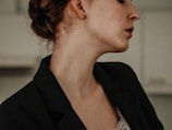 A close-up profile of a person with hair styled in a bun, wearing a dark blazer and lace underneath. The lighting gives a soft, moody atmosphere.