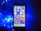A smartphone with a colorful home screen is positioned upright against a dark backdrop. To the left, a cluster of bright blue LED lights casts an illuminating glow.