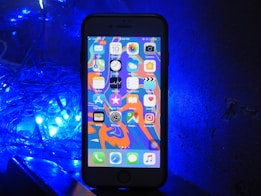A smartphone with a colorful home screen is positioned upright against a dark backdrop. To the left, a cluster of bright blue LED lights casts an illuminating glow.