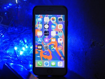 A smartphone with a colorful home screen is positioned upright against a dark backdrop. To the left, a cluster of bright blue LED lights casts an illuminating glow.