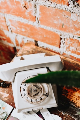 A vintage rotary dial telephone placed on a wooden surface with a background of exposed brick wall. The phone is white and appears slightly worn, suggesting age. A few papers are partially visible beneath the phone.