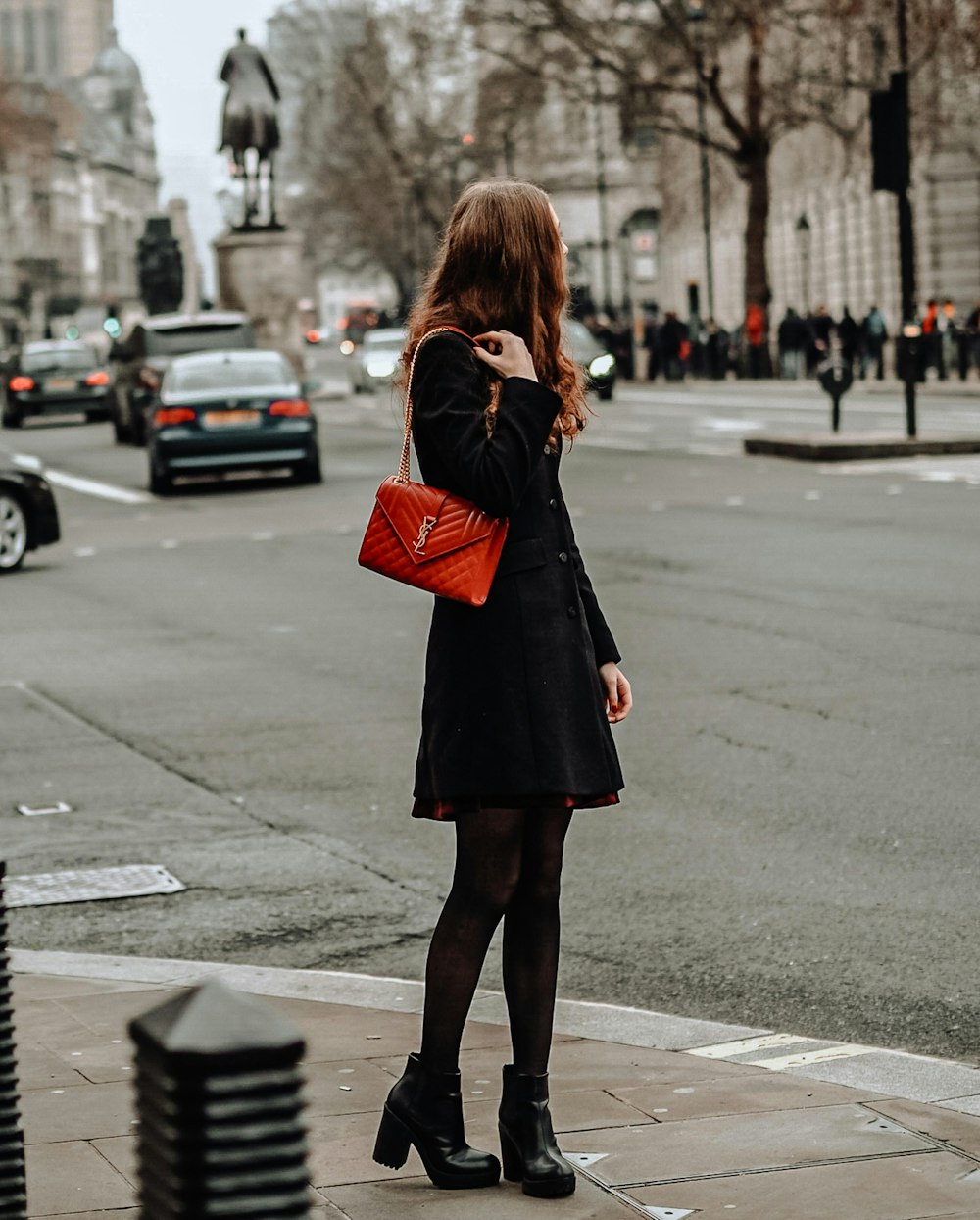 검은 코트와 빨간 가죽 슬링 백을 입은 여자가 낮 동안 보도에 서 있다