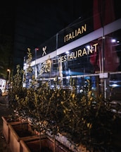 A nighttime scene of an Italian restaurant with a well-lit glass facade. The exterior includes decorative plants in rectangular planters lining the sidewalk. Soft interior and exterior lighting casts a warm glow, highlighting the restaurant signage.
