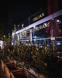 A nighttime scene of an Italian restaurant with a well-lit glass facade. The exterior includes decorative plants in rectangular planters lining the sidewalk. Soft interior and exterior lighting casts a warm glow, highlighting the restaurant signage.