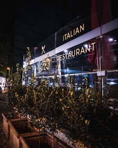 A nighttime scene of an Italian restaurant with a well-lit glass facade. The exterior includes decorative plants in rectangular planters lining the sidewalk. Soft interior and exterior lighting casts a warm glow, highlighting the restaurant signage.