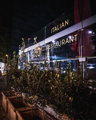 A nighttime scene of an Italian restaurant with a well-lit glass facade. The exterior includes decorative plants in rectangular planters lining the sidewalk. Soft interior and exterior lighting casts a warm glow, highlighting the restaurant signage.