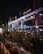 A nighttime scene of an Italian restaurant with a well-lit glass facade. The exterior includes decorative plants in rectangular planters lining the sidewalk. Soft interior and exterior lighting casts a warm glow, highlighting the restaurant signage.