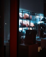A display of leather bags arranged on shelves, illuminated by a blue-tinted light, creating a sleek and stylish atmosphere inside a store. The view is through a glass, showcasing the variety of bags in an organized manner.