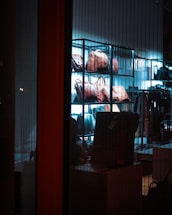 A display of leather bags arranged on shelves, illuminated by a blue-tinted light, creating a sleek and stylish atmosphere inside a store. The view is through a glass, showcasing the variety of bags in an organized manner.