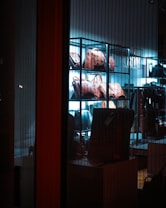 A display of leather bags arranged on shelves, illuminated by a blue-tinted light, creating a sleek and stylish atmosphere inside a store. The view is through a glass, showcasing the variety of bags in an organized manner.