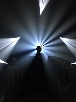 A silhouette of a person wearing headphones stands in front of a DJ setup. Bright, dramatic beams of light radiate outward from behind, creating a dynamic and energetic atmosphere.