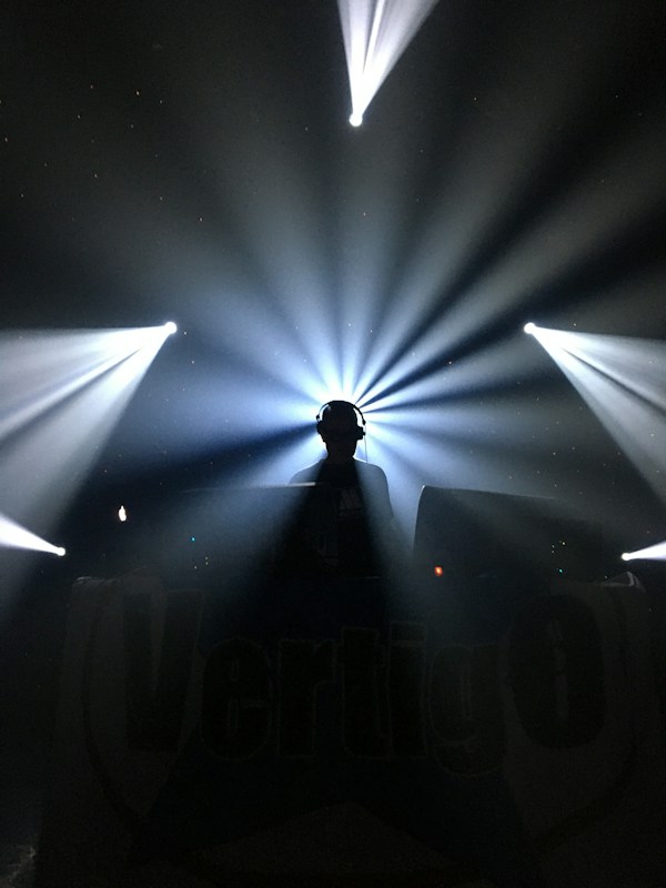 A silhouette of a person wearing headphones stands in front of a DJ setup. Bright, dramatic beams of light radiate outward from behind, creating a dynamic and energetic atmosphere.
