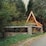 A charming wooden A-frame cabin is nestled among dense, tall pine trees. The cabin features a small, square window at the front, and it is surrounded by a stone and wooden fence. The area is abundant with green foliage and tall yellow wildflowers, enhancing the rustic and secluded atmosphere.