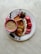 A breakfast arrangement on a decorated plate featuring a pink cup filled with frothy coffee, artisanal bread slices, and sliced strawberries and bananas. The food is artistically placed, highlighting the vibrant colors against a light marble background.