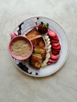 A breakfast arrangement on a decorated plate featuring a pink cup filled with frothy coffee, artisanal bread slices, and sliced strawberries and bananas. The food is artistically placed, highlighting the vibrant colors against a light marble background.
