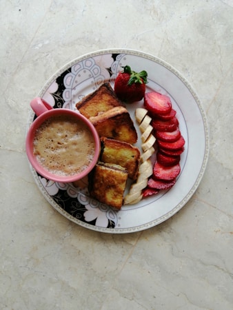 A breakfast arrangement on a decorated plate featuring a pink cup filled with frothy coffee, artisanal bread slices, and sliced strawberries and bananas. The food is artistically placed, highlighting the vibrant colors against a light marble background.
