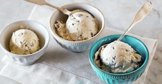 Three bowls of creamy, vanilla ice cream with chocolate chips sit on a light-colored tablecloth. The ice cream is scooped into simple, elegant bowls; one is blue and the others are white or beige. Each bowl has a small spoon with a plain handle, and the setting provides a clean and inviting presentation.