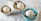 Three bowls of creamy, vanilla ice cream with chocolate chips sit on a light-colored tablecloth. The ice cream is scooped into simple, elegant bowls; one is blue and the others are white or beige. Each bowl has a small spoon with a plain handle, and the setting provides a clean and inviting presentation.
