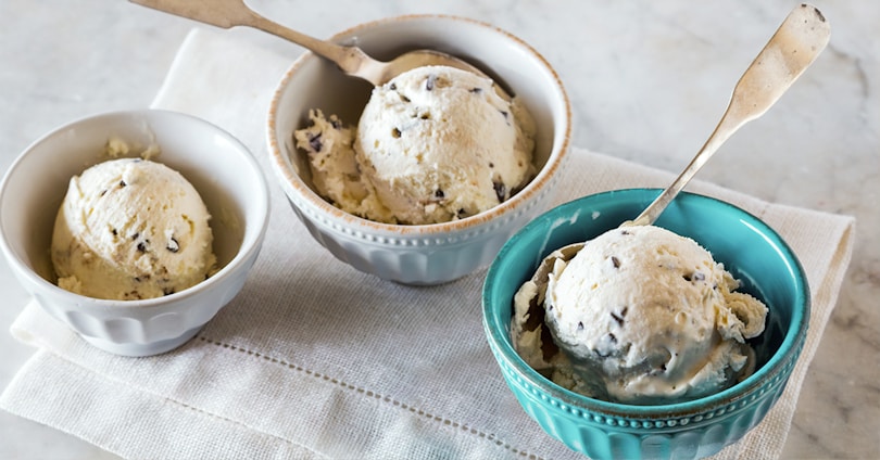 Three bowls of creamy, vanilla ice cream with chocolate chips sit on a light-colored tablecloth. The ice cream is scooped into simple, elegant bowls; one is blue and the others are white or beige. Each bowl has a small spoon with a plain handle, and the setting provides a clean and inviting presentation.