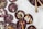Dark chocolate discs decorated with various toppings, including pumpkin seeds, rose petals, coconut flakes, and pink peppercorns, are arranged on a white surface alongside a bowl of melted chocolate with two golden spoons.