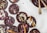 Dark chocolate discs decorated with various toppings, including pumpkin seeds, rose petals, coconut flakes, and pink peppercorns, are arranged on a white surface alongside a bowl of melted chocolate with two golden spoons.