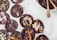 Dark chocolate discs decorated with various toppings, including pumpkin seeds, rose petals, coconut flakes, and pink peppercorns, are arranged on a white surface alongside a bowl of melted chocolate with two golden spoons.