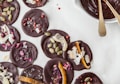 Dark chocolate discs decorated with various toppings, including pumpkin seeds, rose petals, coconut flakes, and pink peppercorns, are arranged on a white surface alongside a bowl of melted chocolate with two golden spoons.