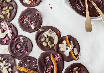 Dark chocolate discs decorated with various toppings, including pumpkin seeds, rose petals, coconut flakes, and pink peppercorns, are arranged on a white surface alongside a bowl of melted chocolate with two golden spoons.