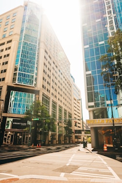An intersection in an urban environment with tall buildings on either side. The sun casts a bright light, illuminating the glass windows of the skyscrapers. Trees line part of the street, and orange traffic cones are visible on the road. A Wells Fargo sign is prominently displayed on one of the buildings.