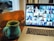 A laptop displays a video conference with multiple participants on the screen, placed on a wooden table next to a ceramic mug. The participants in the video call are visible in small windows, arranged in a grid.