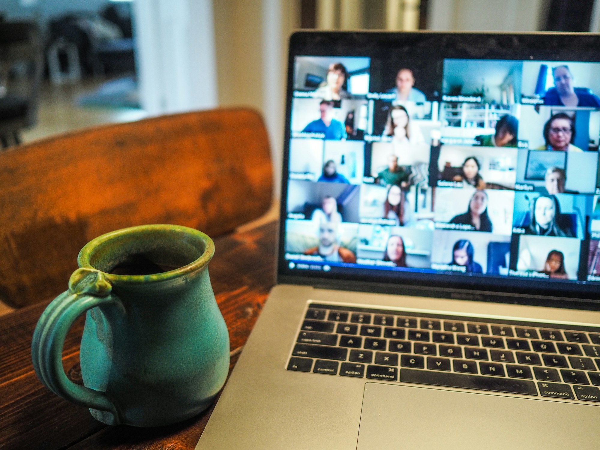 A Macbook Pro displaying a group of people in a video call. The laptop is on a table with a mug beside it.