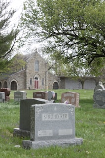 A cemetery with multiple gravestones is set in a grassy area. In the background, an old stone chapel with arched windows and a red door is partially obscured by large, leafy trees. The gravestones vary in shape and size and are arranged throughout the green lawn.