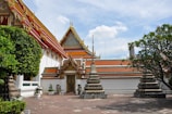 A traditional Thai temple with ornate, decorative roofs and gables highlighted by gold embellishments. The complex exhibits vibrant colors with intricately designed stupas in the foreground set on a paved courtyard. Lush greenery surrounds the temple, complementing the vibrant architecture, under a clear blue sky with scattered clouds.
